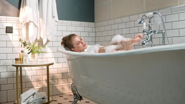 Caucasian attractive young woman taking a bath alone at home washing her leg. Happy pretty female bathing with bubble foam in bathroom. Woman in tub washing body relaxing indoor. Personal hygiene