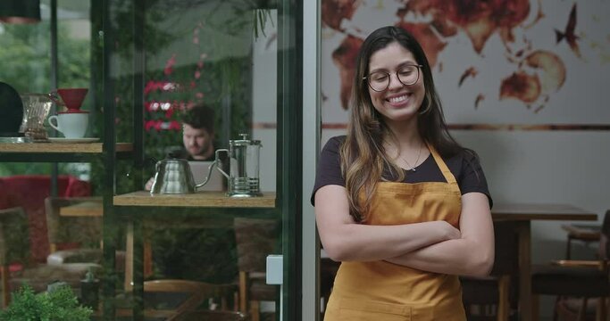 Happy female staff crossing arms smiling at camera in front of coffee shop store wearing yellow apron. A hispanic latin adult girl barista employee of small business restaurant place