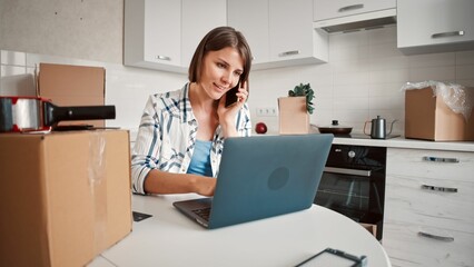 Young female on a laptop and her phone organizing movers surrounded by packed boxes
