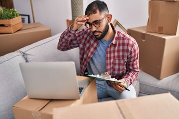 Young hispanic man using laptop reading document at new home