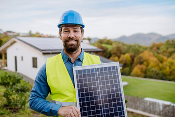 Smiling handyman solar installer standying with solar module while installing solar panel system on...