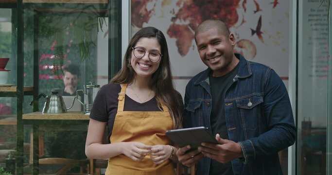 Two diverse employees of small business coffee shop. One black man holding tablet together with female hispanic young woman wearing apron looking at camera smiling. Man and woman staff