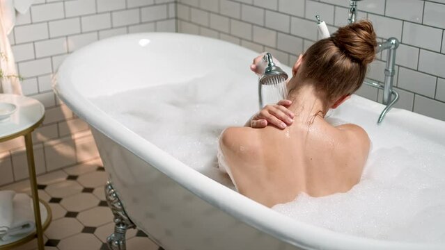 Rear of young adult woman sitting in bathtub naked washing her body holding bath faucet in hand. Back view of female enjoying hot bath with water and soap foam indoors. Spa. Self care and hygiene