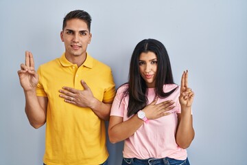 Young couple standing over isolated background smiling swearing with hand on chest and fingers up,...
