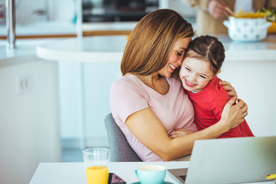 Mother and daughter using laptop and Internet. Freelancer workplace in cozy kitchen. Woman and child girl together. Concept of female business, working mom, freelance, home office. Lifestyle moment.