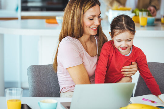 Mother and daughter using laptop and Internet. Freelancer workplace in cozy kitchen. Woman and child girl together. Concept of female business, working mom, freelance, home office. Lifestyle moment.