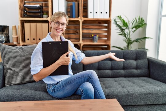 Young blonde woman psychologist holding clipboard sitting on sofa at clinic