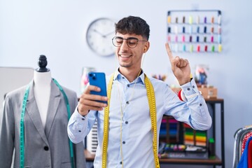 Young hispanic man dressmaker designer using smartphone smiling with an idea or question pointing finger with happy face, number one