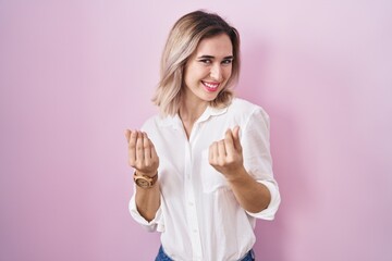 Young beautiful woman standing over pink background doing money gesture with hands, asking for...