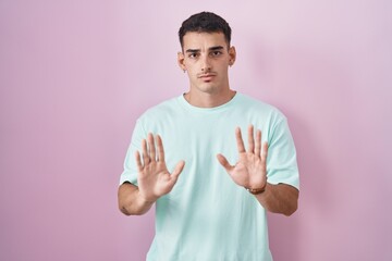 Handsome hispanic man standing over pink background moving away hands palms showing refusal and...