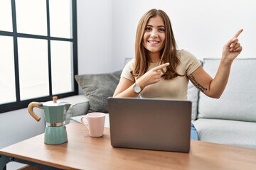 Young brunette woman using laptop at home drinking a cup of coffee smiling and looking at the camera pointing with two hands and fingers to the side.