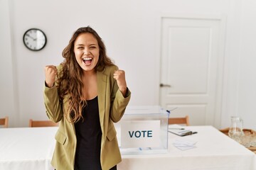 Beautiful hispanic woman standing at political campaign room celebrating surprised and amazed for...