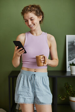 Young Happy Woman Getting Message On Her Smartphone While Drinking Coffee In The Morning In The Room