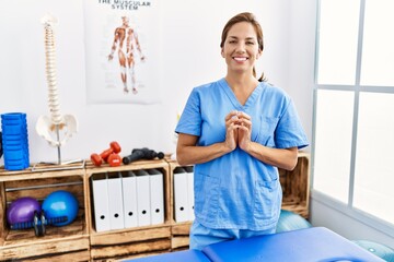 Middle age hispanic physiotherapist woman working at pain recovery clinic hands together and...