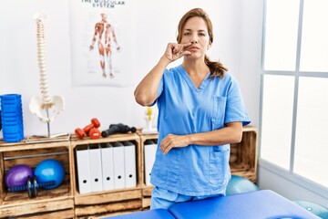 Middle age hispanic physiotherapist woman working at pain recovery clinic mouth and lips shut as...