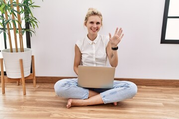 Young blonde woman using computer laptop sitting on the floor at the living room showing and...