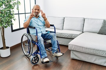 Handsome senior man sitting on wheelchair at the living room gesturing finger crossed smiling with hope and eyes closed. luck and superstitious concept.