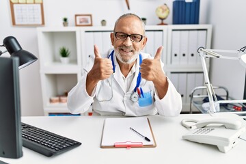 Mature doctor man at the clinic success sign doing positive gesture with hand, thumbs up smiling and happy. cheerful expression and winner gesture.