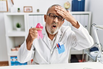 Mature doctor man holding pink cancer ribbon at the clinic stressed and frustrated with hand on head, surprised and angry face