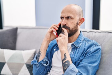 Young bald man talking on smartphone with worried expression at home