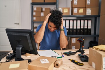 Hispanic man with curly hair working at small business ecommerce suffering from headache desperate and stressed because pain and migraine. hands on head.