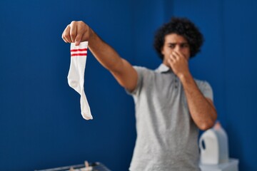 Young hispanic man smelling dirty sock at laundry room