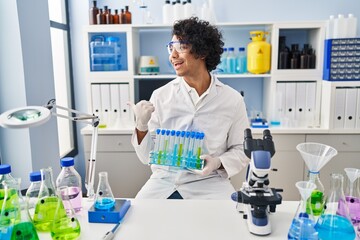Hispanic man with curly hair working at scientist laboratory pointing thumb up to the side smiling...