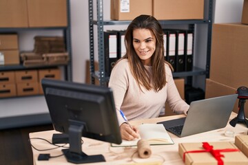 Young beautiful hispanic woman ecommerce business worker using laptop writing on notebook at office