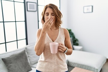 Middle age woman drinking a cup coffee at home bored yawning tired covering mouth with hand....
