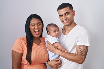 Young hispanic couple with baby standing together over isolated background winking looking at the camera with sexy expression, cheerful and happy face.