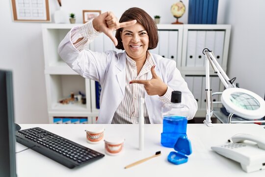 Middle age hispanic dentist woman working at medical clinic smiling making frame with hands and fingers with happy face. creativity and photography concept.