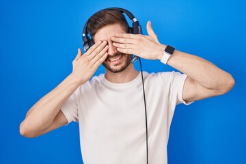 Hispanic man with beard listening to music wearing headphones covering eyes with hands smiling cheerful and funny. blind concept.