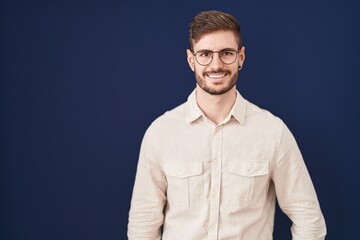Hispanic man with beard standing over blue background winking looking at the camera with sexy expression, cheerful and happy face.