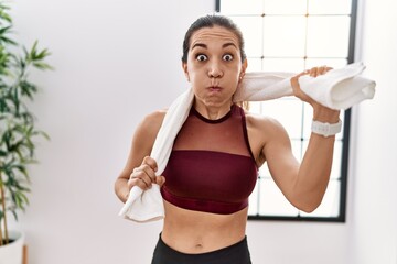 Young hispanic woman wearing sportswear and towel at sport center puffing cheeks with funny face....