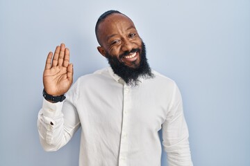 African american man standing over blue background waiving saying hello happy and smiling, friendly welcome gesture