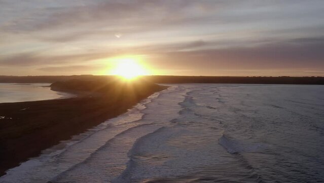 Sand dune beach aerial into colourful sunrise, waves roll onto shore