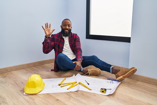 African american man sitting on the floor at new home looking at blueprints showing and pointing up with fingers number five while smiling confident and happy.