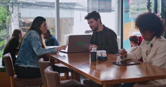 Male colleague showing laptop screen to female friend at coffee shop. Young man friend turning computer to woman sitting at cafe restaurant place. Group of people working remotely