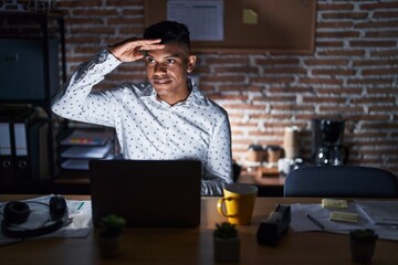 Young hispanic man working at the office at night very happy and smiling looking far away with hand over head. searching concept.