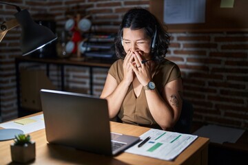 Young hispanic woman working at the office at night laughing and embarrassed giggle covering mouth with hands, gossip and scandal concept