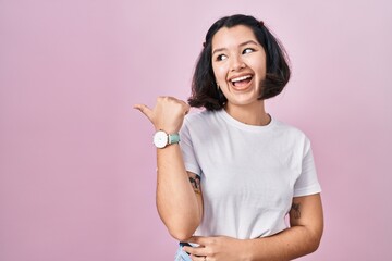 Young hispanic woman wearing casual white t shirt over pink background smiling with happy face looking and pointing to the side with thumb up.