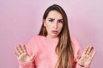 Young hispanic woman standing over pink background moving away hands palms showing refusal and...