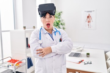 Young hispanic doctor woman wearing doctor uniform and virtual reality glasses in shock face,...