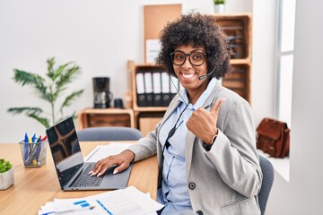 Black woman with curly hair wearing call center agent headset at the office doing happy thumbs up gesture with hand. approving expression looking at the camera showing success.