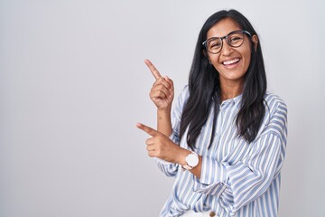Young hispanic woman wearing glasses smiling and looking at the camera pointing with two hands and fingers to the side.