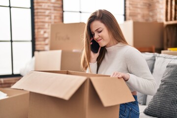 Young blonde woman unboxing package talking on the smartphone at new home