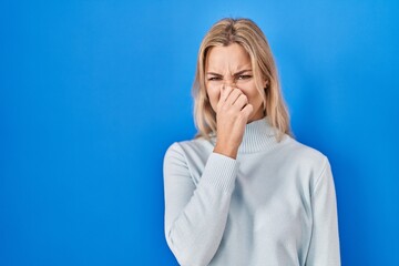 Young caucasian woman standing over blue background smelling something stinky and disgusting,...