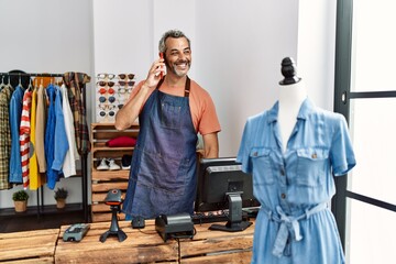 Middle age grey-haired man shop assistant using computer talking on smartphone at clothing store