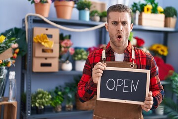 Young caucasian man working at florist holding open sign in shock face, looking skeptical and sarcastic, surprised with open mouth