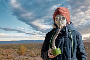 A boy in a gas mask and warm clothes on the background of the desert on a sunny day. In his hand he holds a gas mask with a filter. Blue sky with dramatic clouds. Apocalypse concept